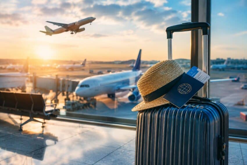 Travel essentials — a suitcase, straw hat and passport resting against an airport window at sunset, with planes on the tarmac beyond